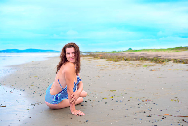 Jonge brunette meisje in gestreepte zwembroek zittend op het strand op het zand. Felle zon blauwe hemel wolken schijnt - Foto, afbeelding