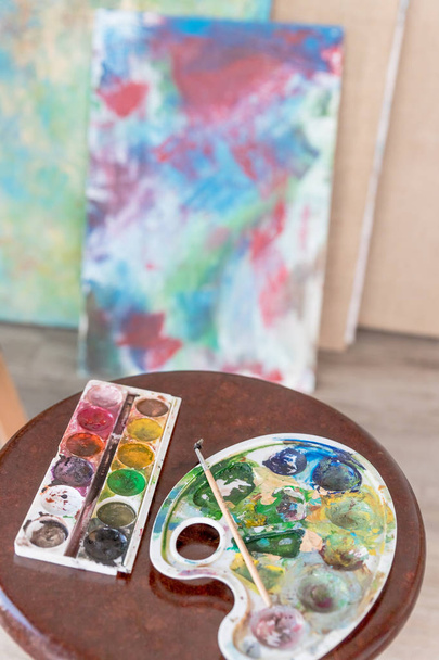 Artistic equipment in studio, easel, paintbrushes, tubes of paint, palette and paintings on work artist table.painting picture on a canvas.art school, creativity concept - Photo, Image