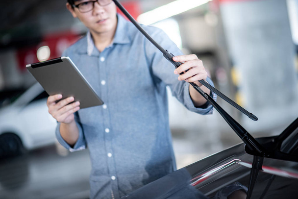 Nuori aasialainen automekaanikko, jolla on digitaalinen tabletti, joka tarkistaa tuulilasinpyyhkimen autotallissa. Koneenrakennusinsinööri, joka työskentelee autoteollisuudessa. Autojen huolto ja korjaus
 - Valokuva, kuva