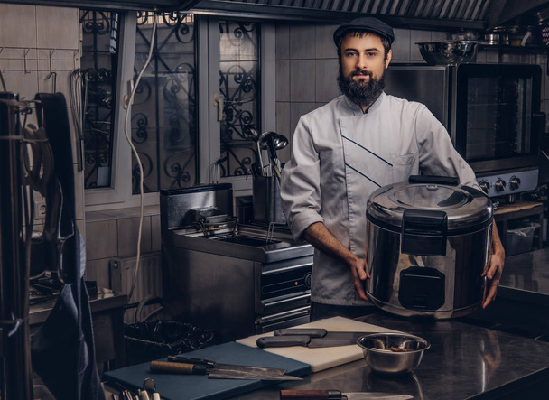 Bearded cook dressed in uniform and hat holds big multicooker in kitchen. - Photo, Image