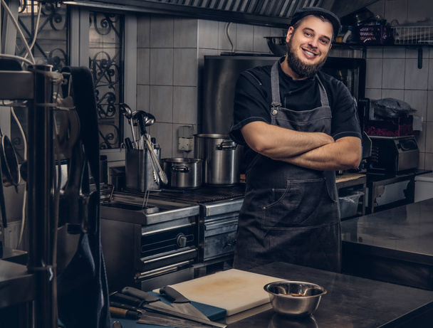 Χαμογελώντας γενειοφόρος μάγειρας στην μαύρη στολή στέκεται με σταυρωμένα τα χέρια στην κουζίνα. - Φωτογραφία, εικόνα