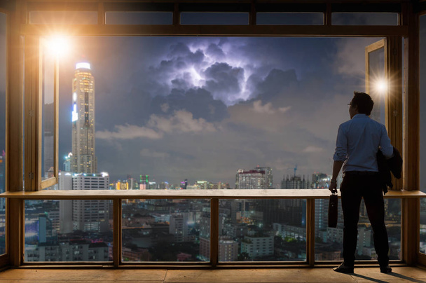 Concept visión, Joven hombre de negocios con chaqueta cómoda traje casual de pie sosteniendo una bolsa de trabajo en la sala de trabajo cerca de la ventana de vidrio y mirando a través de los edificios modernos en el horizonte
. - Foto, imagen