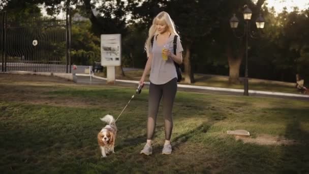Front view of beautiful smilling woman with long blonde hair walking with her dog in the park .She is wearing casual clothes and holding drink in hand - Filmati, video
