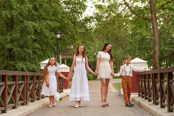 young mother with children siblings in white dresses walking along the bridge holding hands and laughing smiling - Фото, изображение