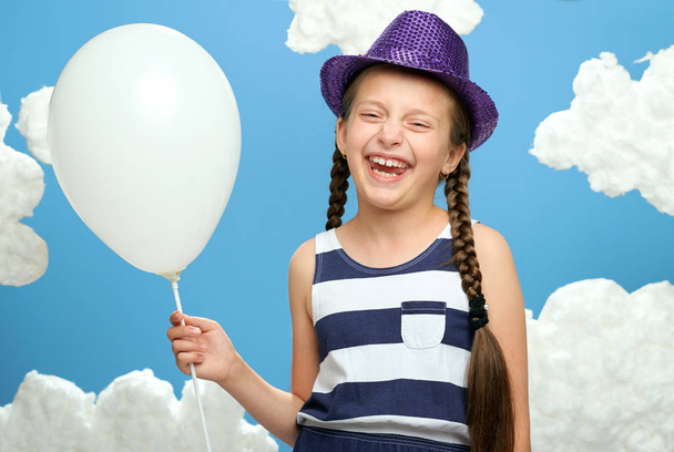 girl dressed in striped dress and color hat posing on a blue background with cotton clouds, white air balloon, the concept of summer, happiness and holiday - Foto, afbeelding