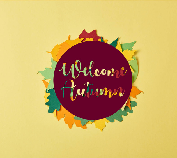 flat lay with colorful papercrafted foliage with "welcome autumn" lettering in circle on yellow background - Photo, Image