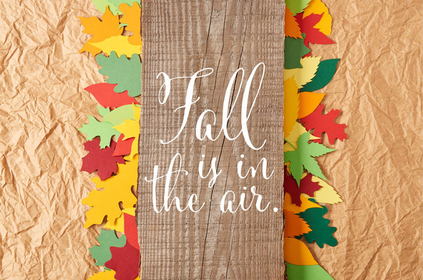 top view of wooden plank with "fall is in the air" inspiration and colorful paper leaves arrangement on crumpled paper backdrop - Fotografie, Obrázek