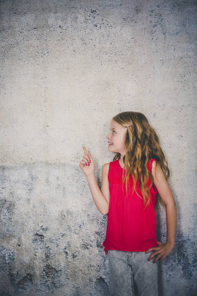 beautiful girl in red shirt pointing aside while standing in front of concrete background  - 写真・画像