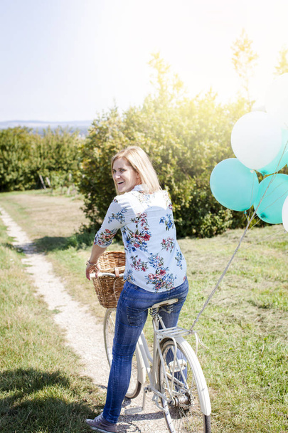 Onnellinen nainen ratsastus polkupyörällä ilmapalloilla kesäniityllä aurinkoisena päivänä
 - Valokuva, kuva