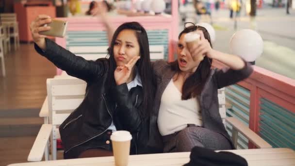Ragazze spensierate stanno prendendo selfie con prendere bevande seduti in caffè insieme e utilizzando smartphone. Giovani donne sono in posa, occhiali tintinnanti e mostrando gesti delle mani
. - Filmati, video