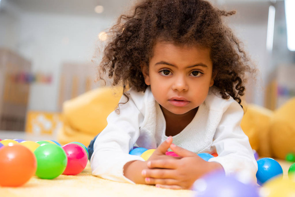 adorable afroamericano niño acostado en la alfombra con juguetes en el jardín de infantes y mirando a la cámara
 - Foto, Imagen