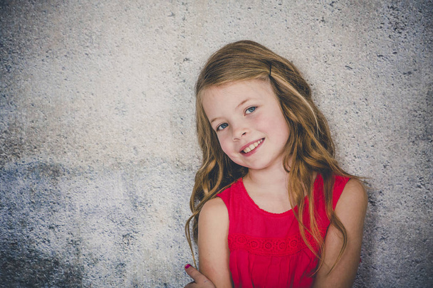 happy attractive little girl with blond curly hair smiling and looking at camera while posing in front of concrete background  - Фото, зображення