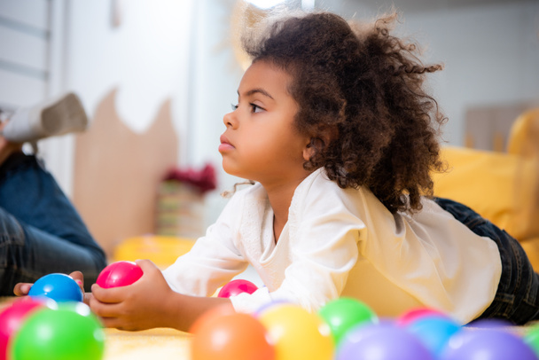 side view of adorable african american kid lying on carpet with toys and looking away in kindergarten - Foto, Imagem