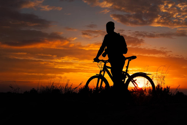 美しい夕日に牧草地でサイクリング スポーツ人のシルエット。カメラと自転車の若い男. - 写真・画像