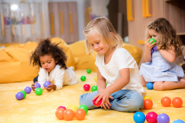 adorable multicultural kids playing with colored balls on floor in kindergarten - Foto, imagen