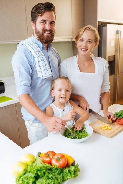 Young family of three cooking healthy salad for lunch in kitchen and looking at camera  - Photo, Image