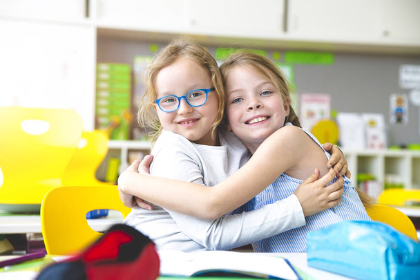 two schoolgirls embracing and showing thumbs up at classroom - Φωτογραφία, εικόνα