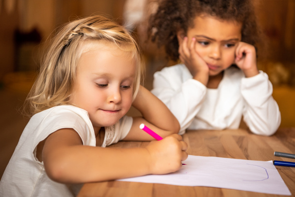 criança afro-americana entediada olhando para longe, criança branca desenhando à mesa no jardim de infância
 - Foto, Imagem