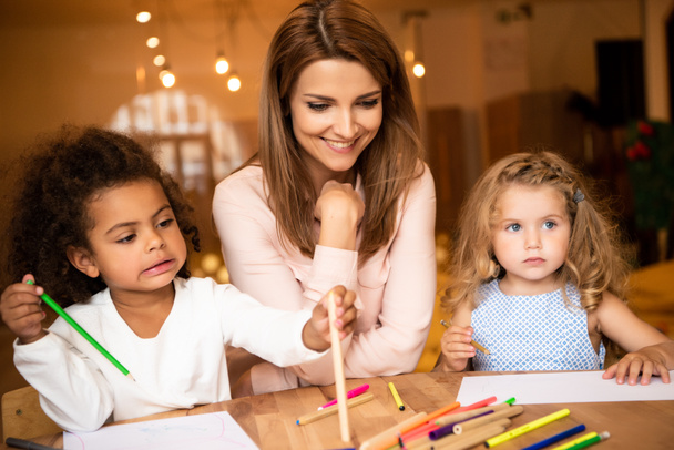 cheerful educator sitting near multicultural kids drawing in kindergarten - Photo, Image