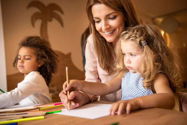 счастливый репетитор помогает ребенку рисовать в детском саду
 - Фото, изображение