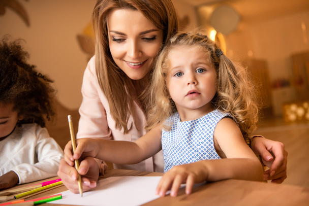 cheerful educator helping adorable kid drawing in kindergarten - Photo, Image