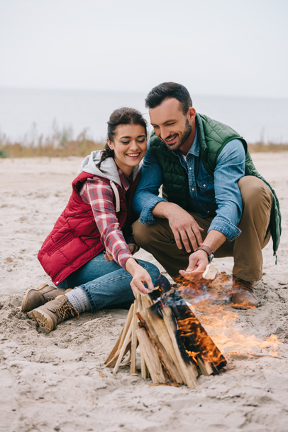 couple roasting marshmallow on campfire on sandy beach - 写真・画像
