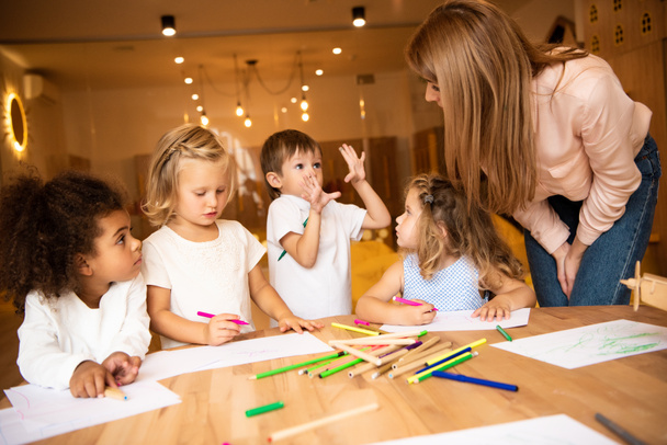 мультиэтнические дети рисуют в детском саду, педагог смотрит на жесты мальчика
 - Фото, изображение