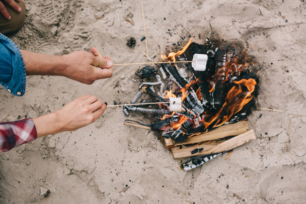 砂浜のビーチでキャンプファイヤーでマシュマロを焙煎のカップルのショットをトリミング - 写真・画像