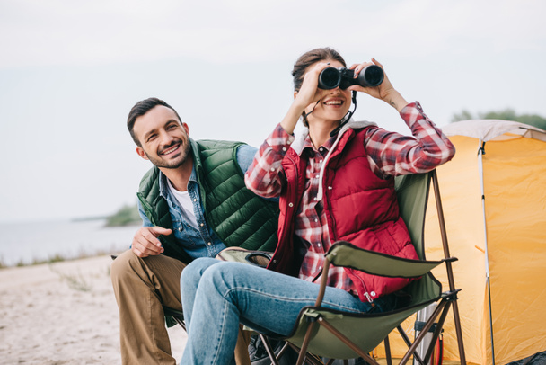 couple with binoculars resting on chairs on camping - Photo, Image