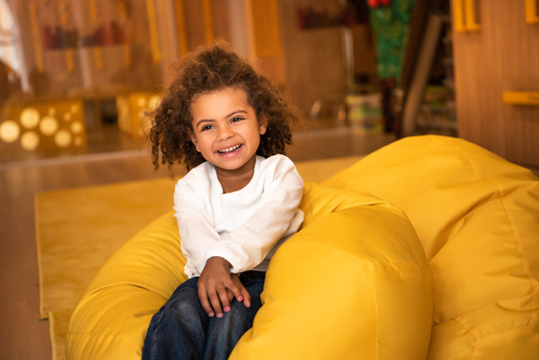 smiling adorable african american kid sitting on bean bag chair in kindergarten - Photo, Image