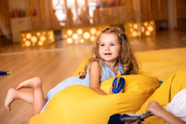 smiling adorable kid lying on bean bag chair with toy and looking away in kindergarten - Photo, Image