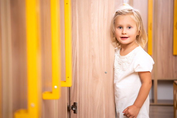 adorable kid stojący w pobliżu open locker i patrząc na kamery w szatni przedszkola - Zdjęcie, obraz