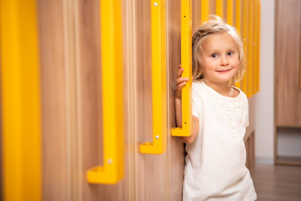 cheerful adorable kid standing in kindergarten cloakroom and looking at camera - Foto, imagen