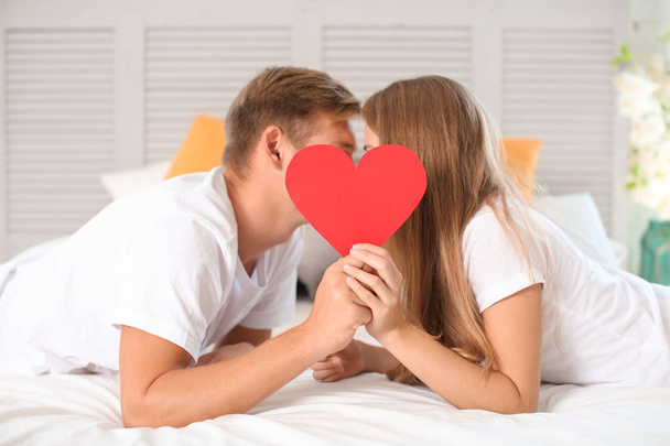 Happy young couple hiding faces behind red heart while lying on bed at home - Zdjęcie, obraz