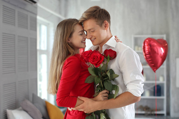 Feliz pareja joven con rosas rojas abrazándose en casa
 - Foto, imagen