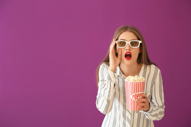 Emotional young woman with cup of popcorn and 3D cinema glasses on color background - Foto, Imagen