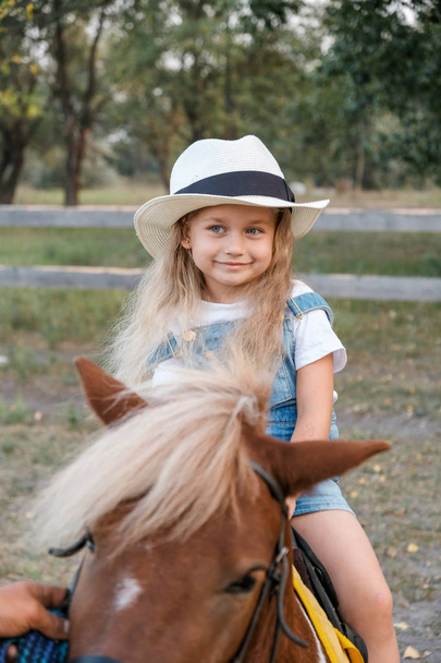 A cute little blonde girl is sitting on a pony in autumn. - Фото, зображення