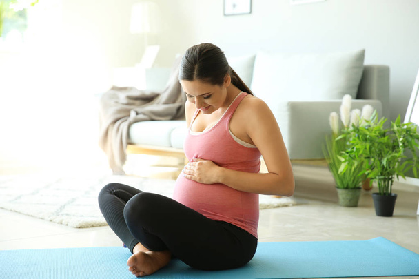 Νεαρή έγκυος γυναίκα κάθεται στο χαλάκι γιόγκα στο σπίτι - Φωτογραφία, εικόνα