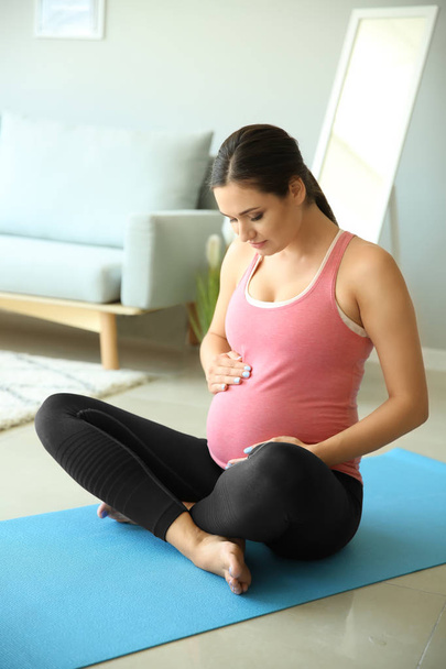 Mujer embarazada joven sentada en la esterilla de yoga en casa
 - Foto, Imagen