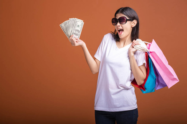 Woman holding shopping bag and cash isolated in orange background. Young asian woman in white t-shirt, sun glasses in very happy mood. Holiday shopping concept. - Photo, image