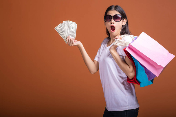 Vrouw met boodschappentas en cash geïsoleerd in oranje achtergrond. Jonge Aziatische vrouw in wit t-shirt, zon brillen in zeer verrassing stemming. Holiday shopping concept. - Foto, afbeelding