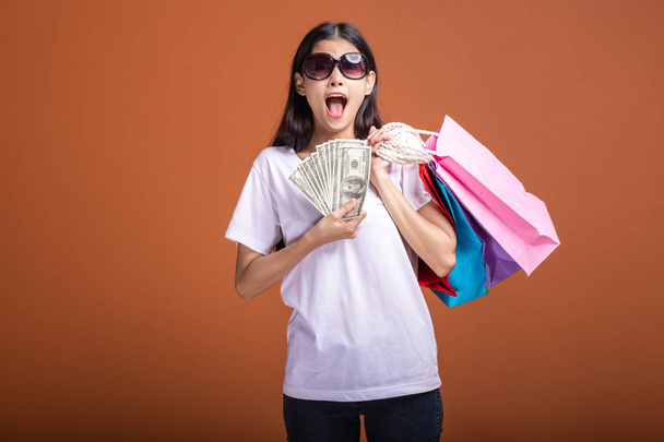 ショッピング バッグとオレンジ色の背景に分離されたキャッシュを保持している女性。白い t シャツの若いアジア女性、太陽メガネ機嫌が非常に興奮しています。休日ショッピング概念. - 写真・画像