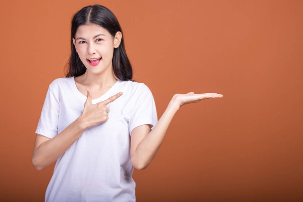 Woman pointing finger with open palm over orange background. Young Asian hipster woman in white t-shirt posing open palm with happy emotion. - Photo, image
