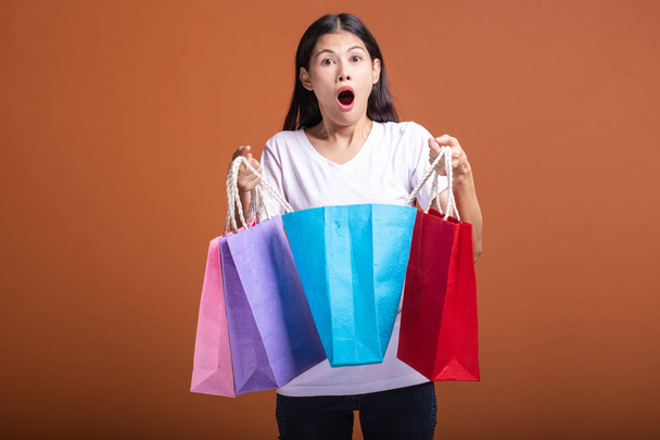 Woman holding shopping bag isolated in orange background. Young asian woman in white t-shirt, very shock mood, looking into bag. Shopping concept. - Foto, imagen