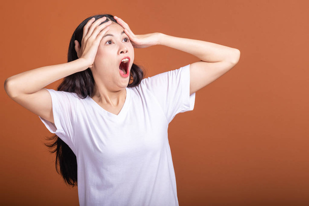 Surprise woman over orange background. Young Asian hipster woman in white t-shirt with very shock pose, hands cover head. Young woman hipster concept. - Foto, Imagen