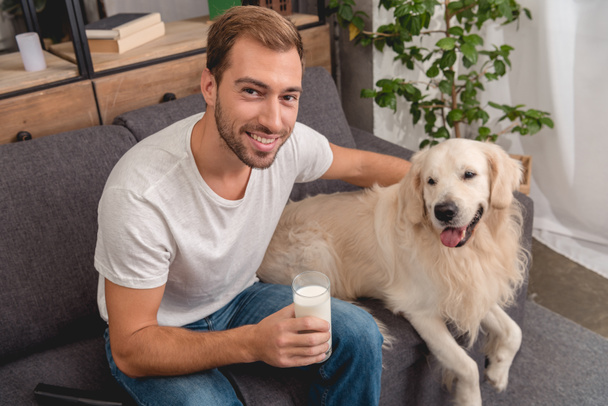 vysoký úhel pohled usmívající se mladý muž s sklenici mléka a rozkošný zlatý retrívr pes při pohledu na fotoaparát - Fotografie, Obrázek
