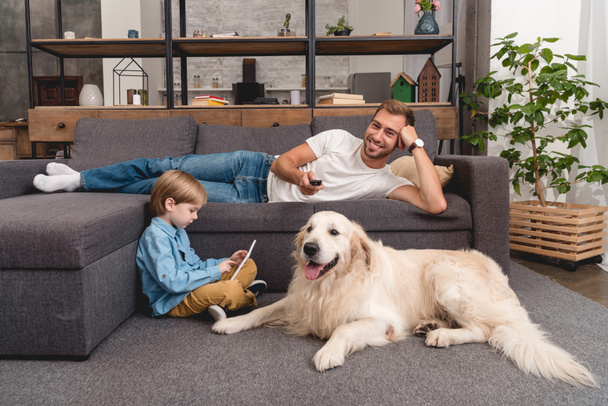happy father watching tv on couch while his son using tablet on floor with golden retriever - Fotoğraf, Görsel