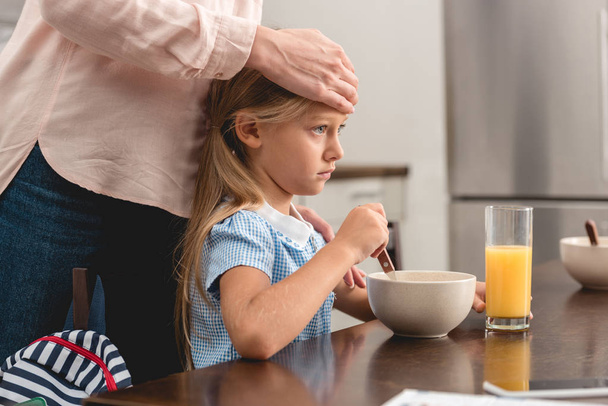oříznuté shot matky kontrola teploty nemocné dcery s rukou, zatímco ona snídani - Fotografie, Obrázek