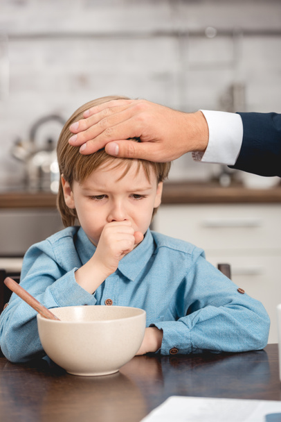 cropped shot of father checking temperature of sick son with hand during breakfast - Photo, Image