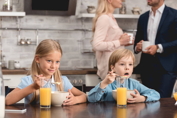 cute little kids having breakfast together at kitchen while parents drinking coffee on background - Zdjęcie, obraz